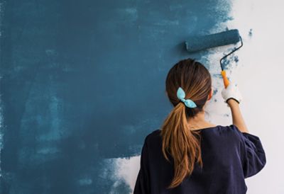 A woman using a paint roller applies dark blue paint to a wall. 