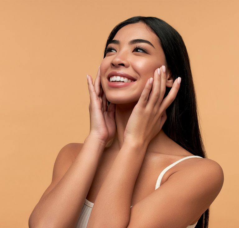 A smiling person with glowing skin touches their cheeks with both hands.  