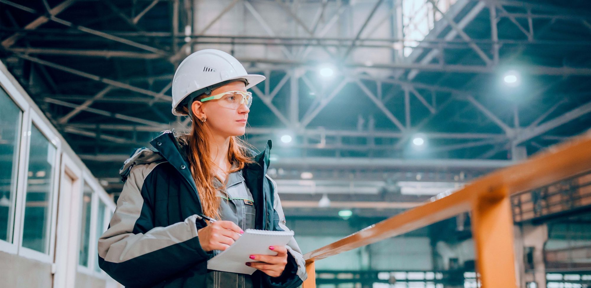 An employee wearing a helmet and safety glasses takes notes while monitoring manufacturing. 