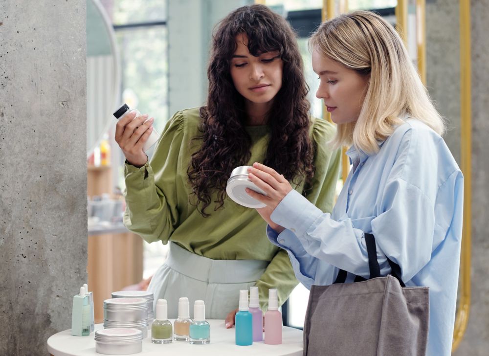Two people look at a display of cosmetics packaging and each hold an item. 