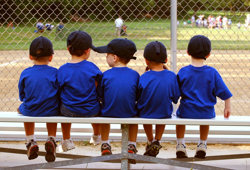 Five little boys in matching shirts and hats sit on a bleacher at a ball field. 