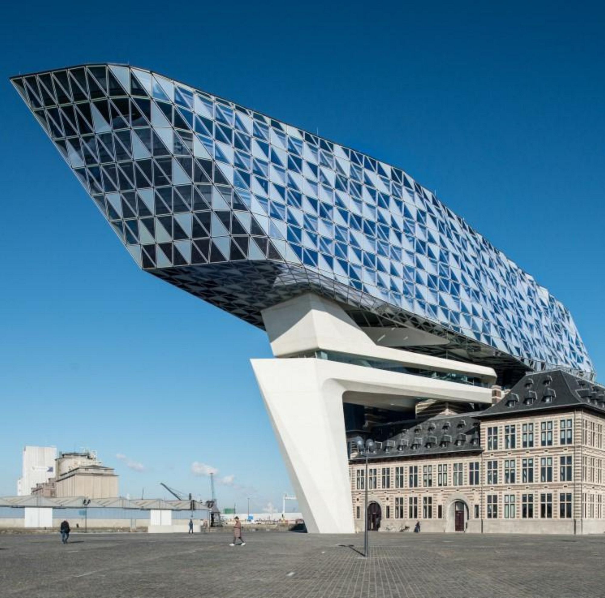 A modern,  glass building with a geometric facade rises above a historic square under a clear blue sky. 
